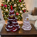 Two glass Christmas trees, one red and one clear with gold accents, on a table in front of a decorated Christmas tree.