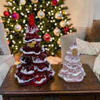 Two glass Christmas trees, one red and one clear with gold accents, on a table in front of a decorated Christmas tree.