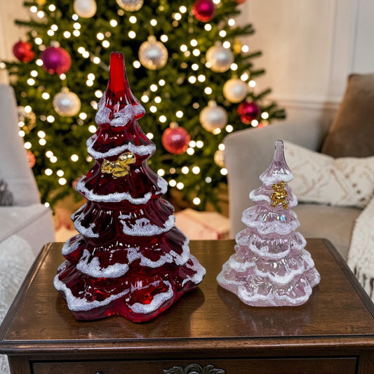Two glass Christmas trees, one red and one clear with gold accents, on a table in front of a decorated Christmas tree.